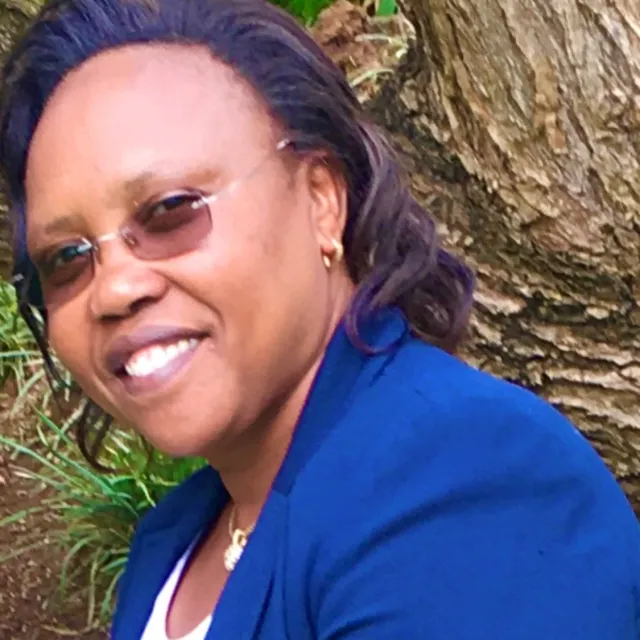Joyce wearing glasses and a blue blazer and smiling beside a tree