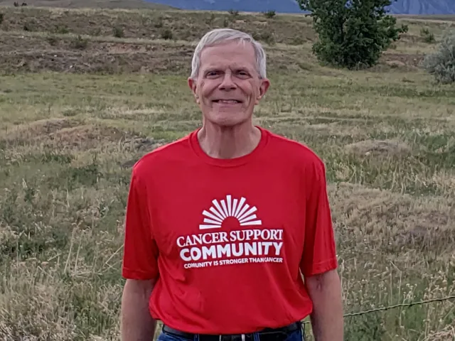 Paul smiles in a red Cancer Support Community T-shirt with the foothills of the Colorado Rockies behind him