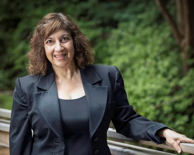Kerri Susko wears a black business suit and smiles from a wooden bridge beside trees