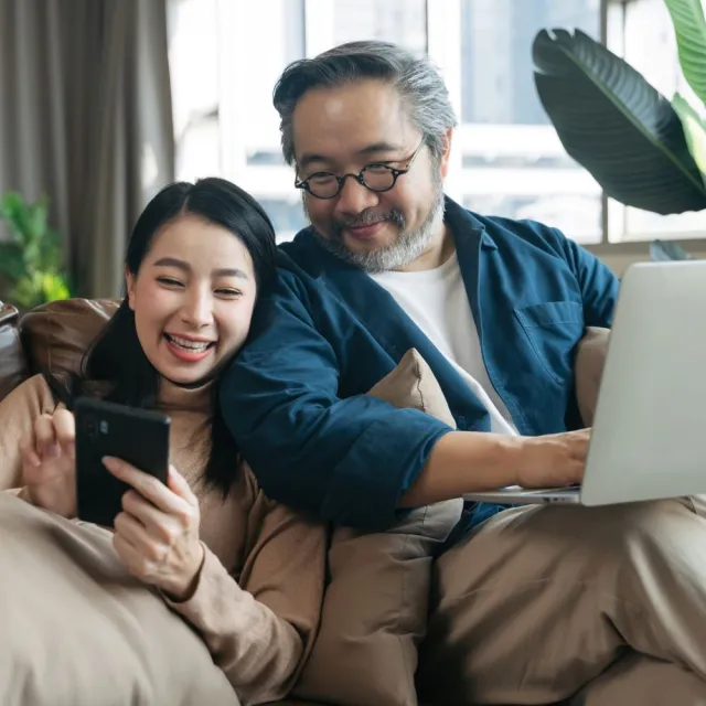 A couple smiles while sitting together on their couch viewing their phone and laptop
