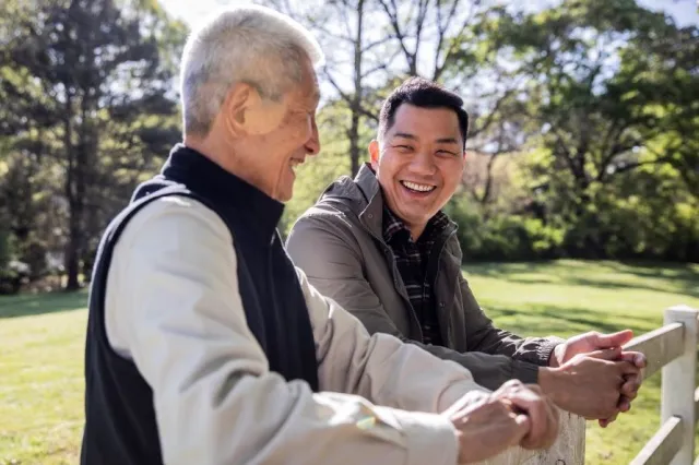 A senior man in a jacket vest laughs with his son outdoors where they rest their hands on a fence