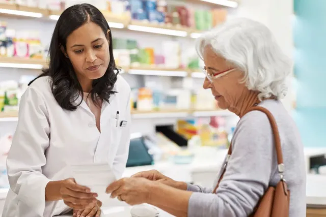 A senior woman stands at a pharmacy counter showing a female pharmacist her prescription