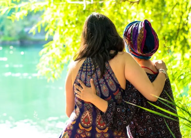 A woman wearing a colorful head scarf embraces her sister along the bank of a sunlit river