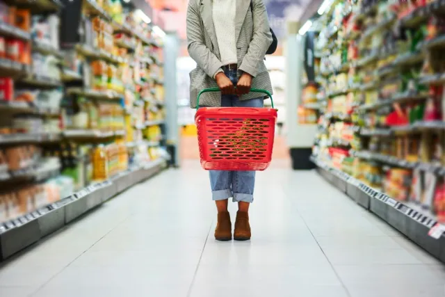 A woman carrying a red grocery basket stands in the middle of a supermarket aisle