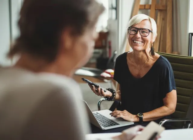A woman with short gray hair and dark glasses smiles with a phone and laptop while talking with an unseen woman