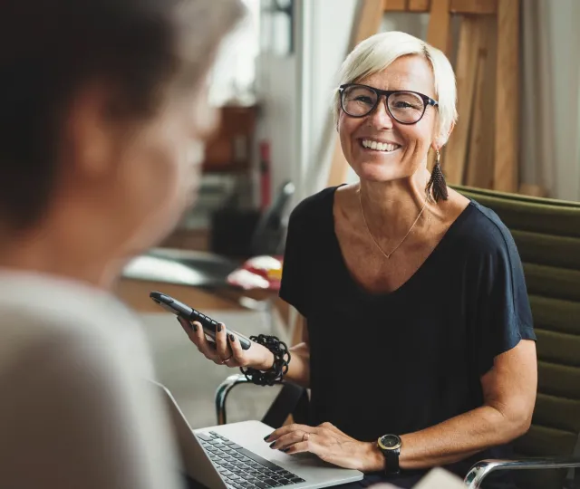 A woman smiles at an unseen person while sitting in a chair and holding a smartphone and a laptop