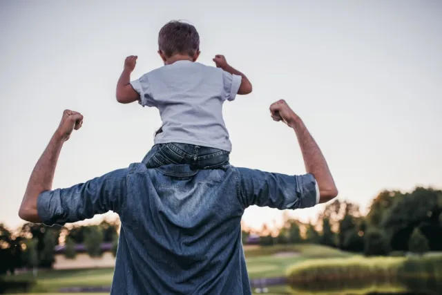 A young father carries his toddler son piggyback as they both raise their fists toward the sky