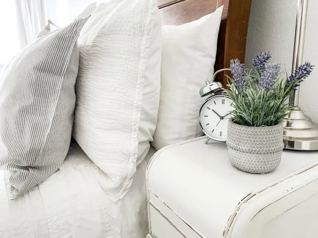 Pillows are stacked on a bed next to a table with alarm clock and potted lavender flowers