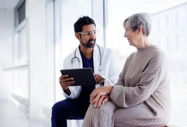 A woman with short gray hair talks with a doctor in a lab coat in a brightly lit room