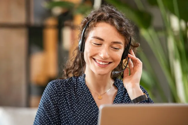 A smiling woman talks to a customer on a headset while looking at her laptop
