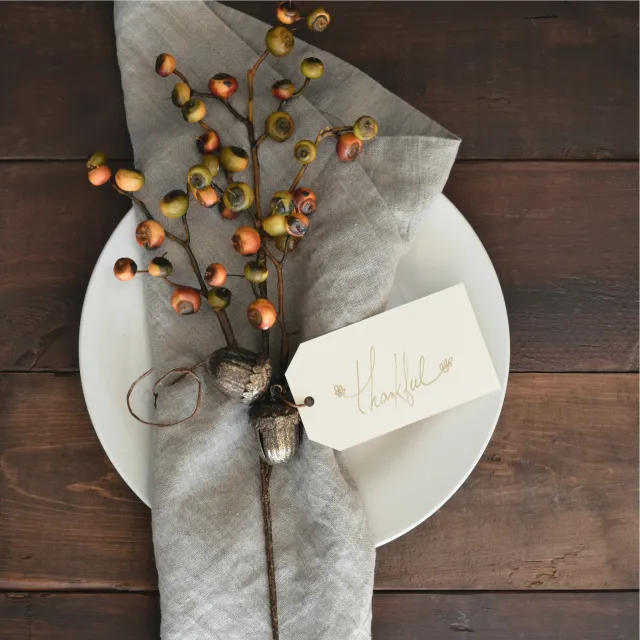 A dinner napkin rests on a small plate with dried berries, decorative silver acorns, and a tag with the handwritten word Thankful
