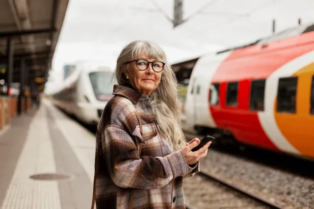 A mature woman with long hair and dark glasses waits at a train station