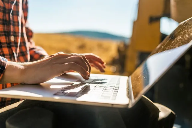 Unseen person wearing a flannel shirt types on a laptop outdoors in a mountain landscape