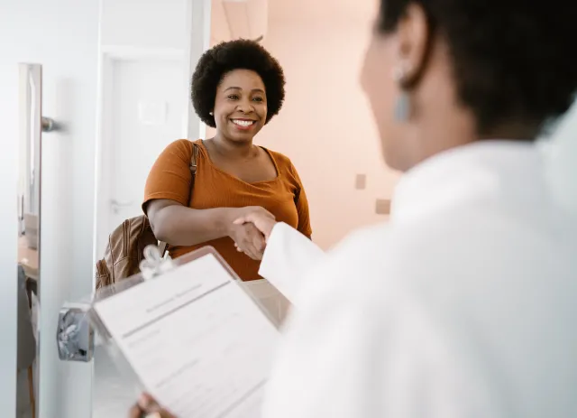 A woman arriving at a medical office smiles and shakes hands with a female doctor