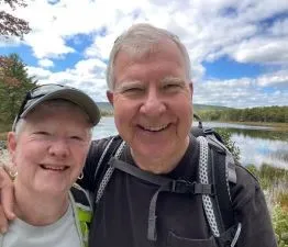Lyn and her husband smile from a hiking trail near a lake that reflects the blue sky above