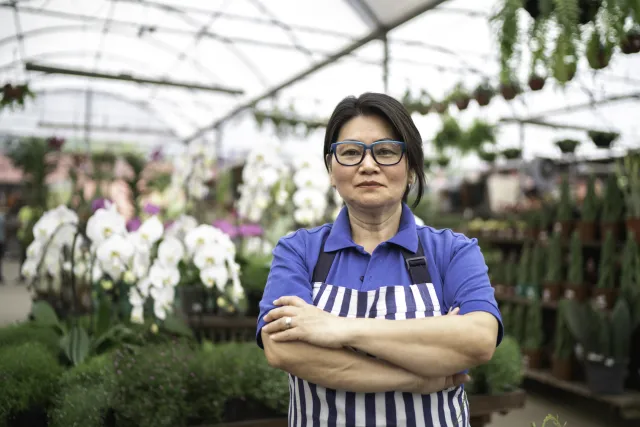 A smiling flower shop owner wearing a striped apron stands amid flowers and plants 