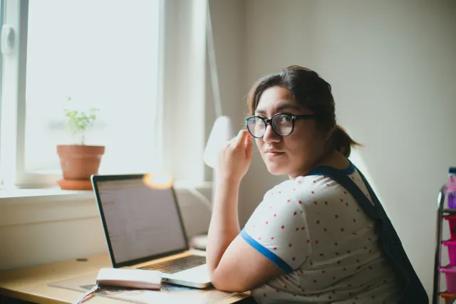 A young woman wearing glasses and seated at a desk looks up from her laptop