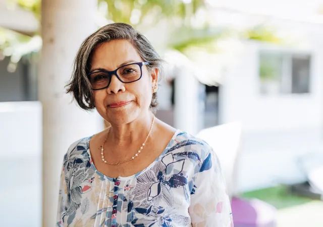 A woman wearing glasses smiles in a friendly manner from a sunlit porch