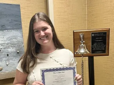 Abbie, smiling, holds a treatment completion certificate beside a bell in her cancer center