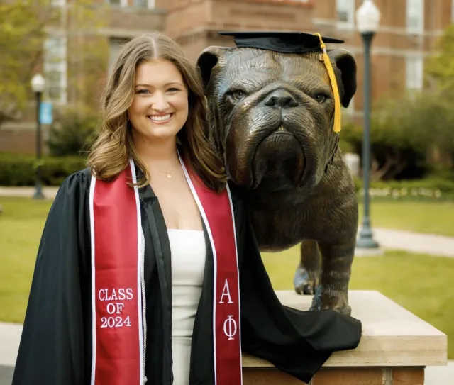 Abbie smiles in a black graduation gown, standing outside on her college campus