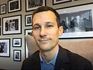 White man smiling softly in front of a wall full of picture frames