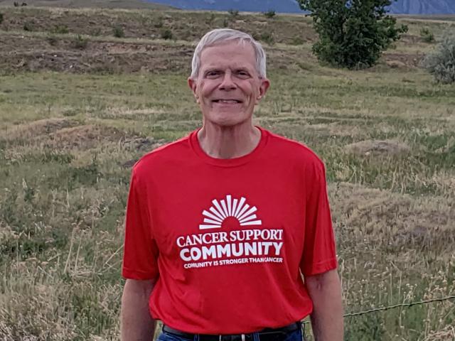 Paul smiles in a red Cancer Support Community T-shirt with the foothills of the Colorado Rockies behind him