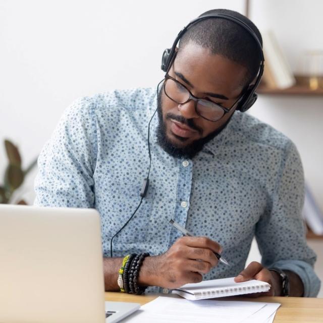 A man wearing headphones views his laptop screen and takes notes with a pen in his hand