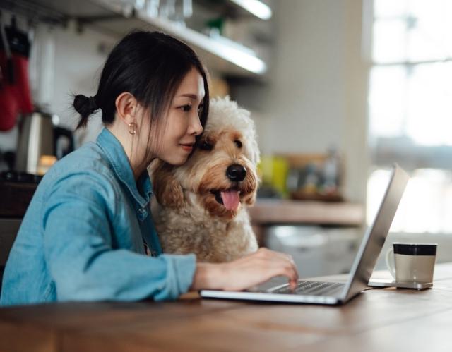 A young woman smiles while viewing her laptop on a kitchen table with a dog sitting beside her