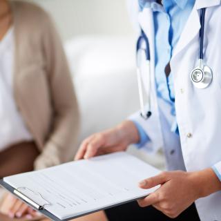 A doctor in a white coat holds a clipboard while talking with a patient