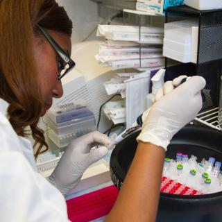 A medical researcher wearing a white lab coat and gloves examines a test vial in a research lab
