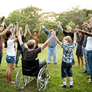 A group of people reflecting diverse ages and races stands in a circle, clasps one another's hands in unity and raises them to the sky