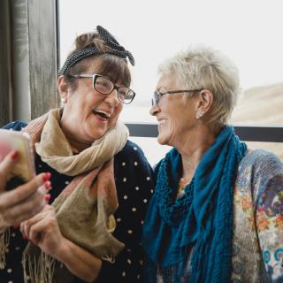 Two women friends laugh together beside a window while looking at their smartphone