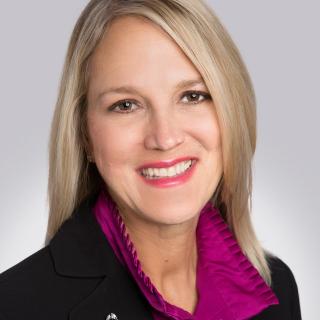 A headshot of CSC's new CEO Debbie Weir shows her smiling and wearing a purple blouse and black blazer with a cancer awareness ribbon lapel pin