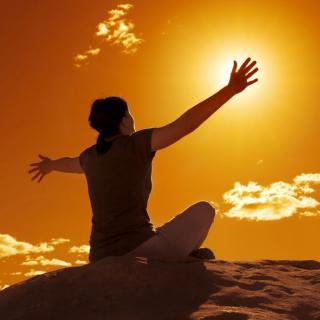 A woman sitting on a rock extends her arms out as if embracing the setting sun in an orange-lit sky
