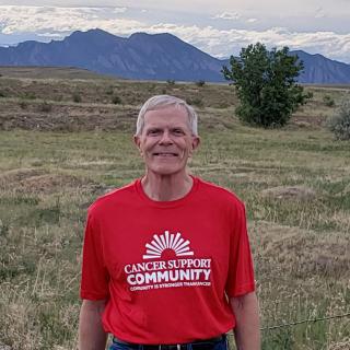 Dressed in jeans and a red Cancer Support Community T-shirt, Paul smiles outside with the Boulder foothills behind him