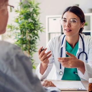 A woman doctor in a white lab coat speaks with a man sitting at a desk in a doctor's office