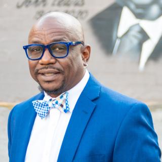 Tony Walker wears a blue and white bow tie, blue blazer, white blouse, and glasses with blue frames