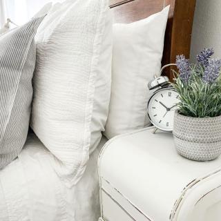 Pillows are stacked on a bed next to a table with alarm clock and potted lavender flowers