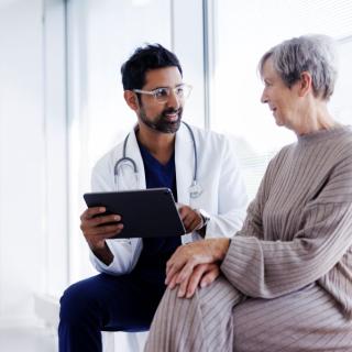 A woman with short gray hair talks with a doctor in a lab coat in a brightly lit room