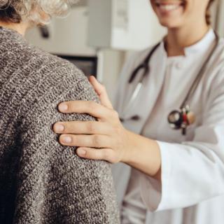 A female doctor in a white lab coat rests a comforting hand on a patient's shoulder