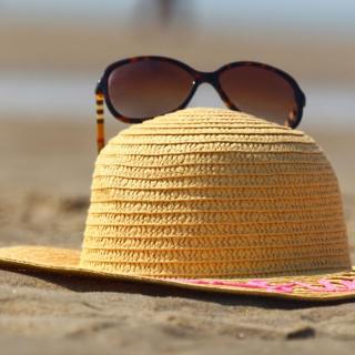 A pair of brown sunglasses sits on a straw sun hat resting on a sandy beach