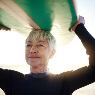 A senior woman with short gray hair holds a surf board over her head on a sunny beach