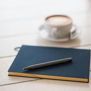 A black pen rests on a hard-cover notebook beside a cup of coffee
