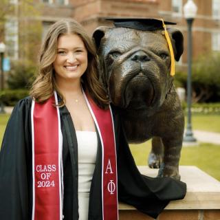 Abbie smiles in a black graduation gown, standing outside on her college campus