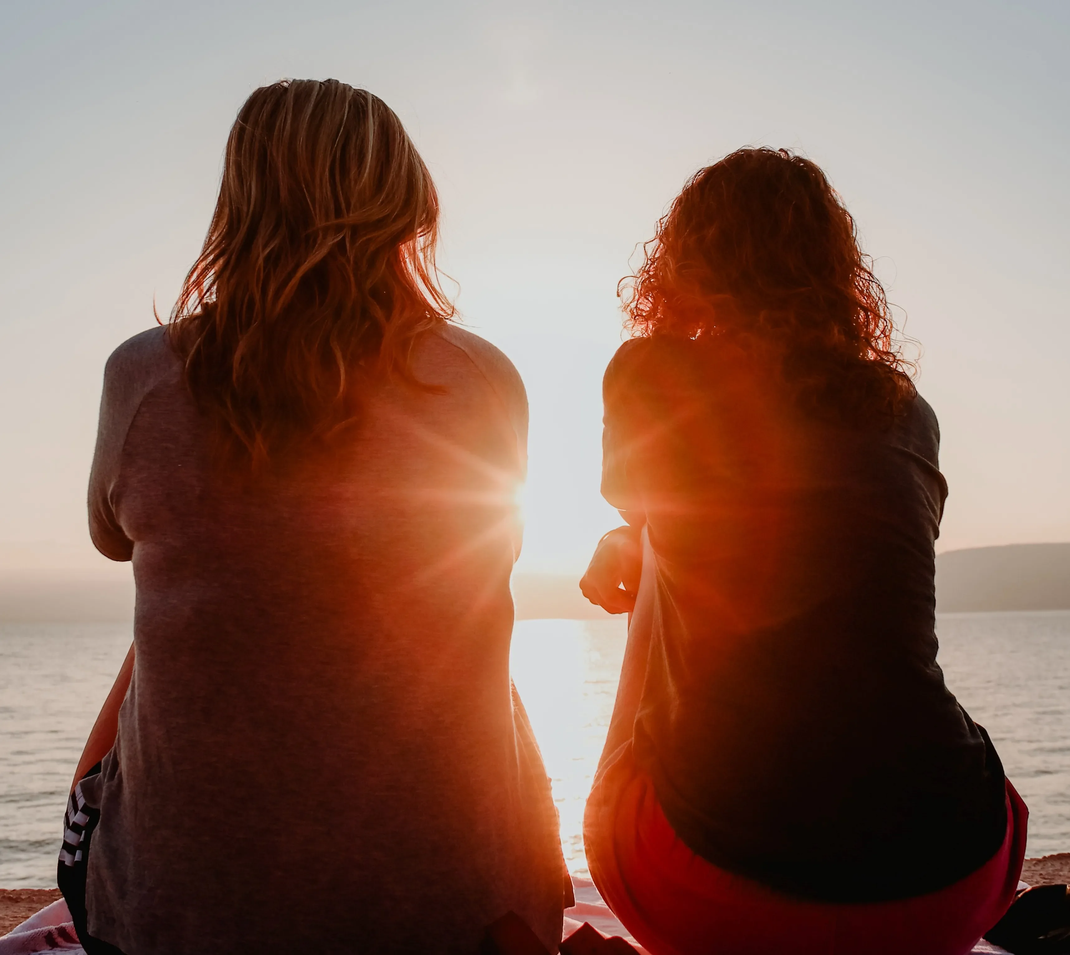 Two women friends sit together looking out at a lake as the sun sets