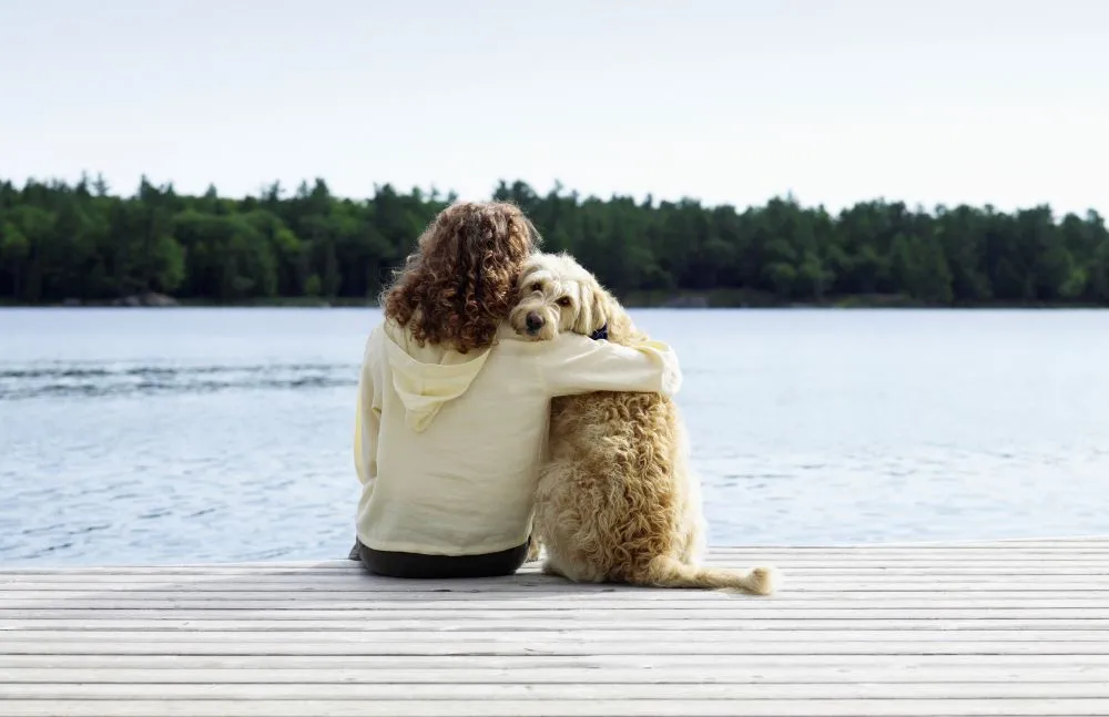 A young woman hugs her dog as they sit together on the edge of a pier overlooking a lake