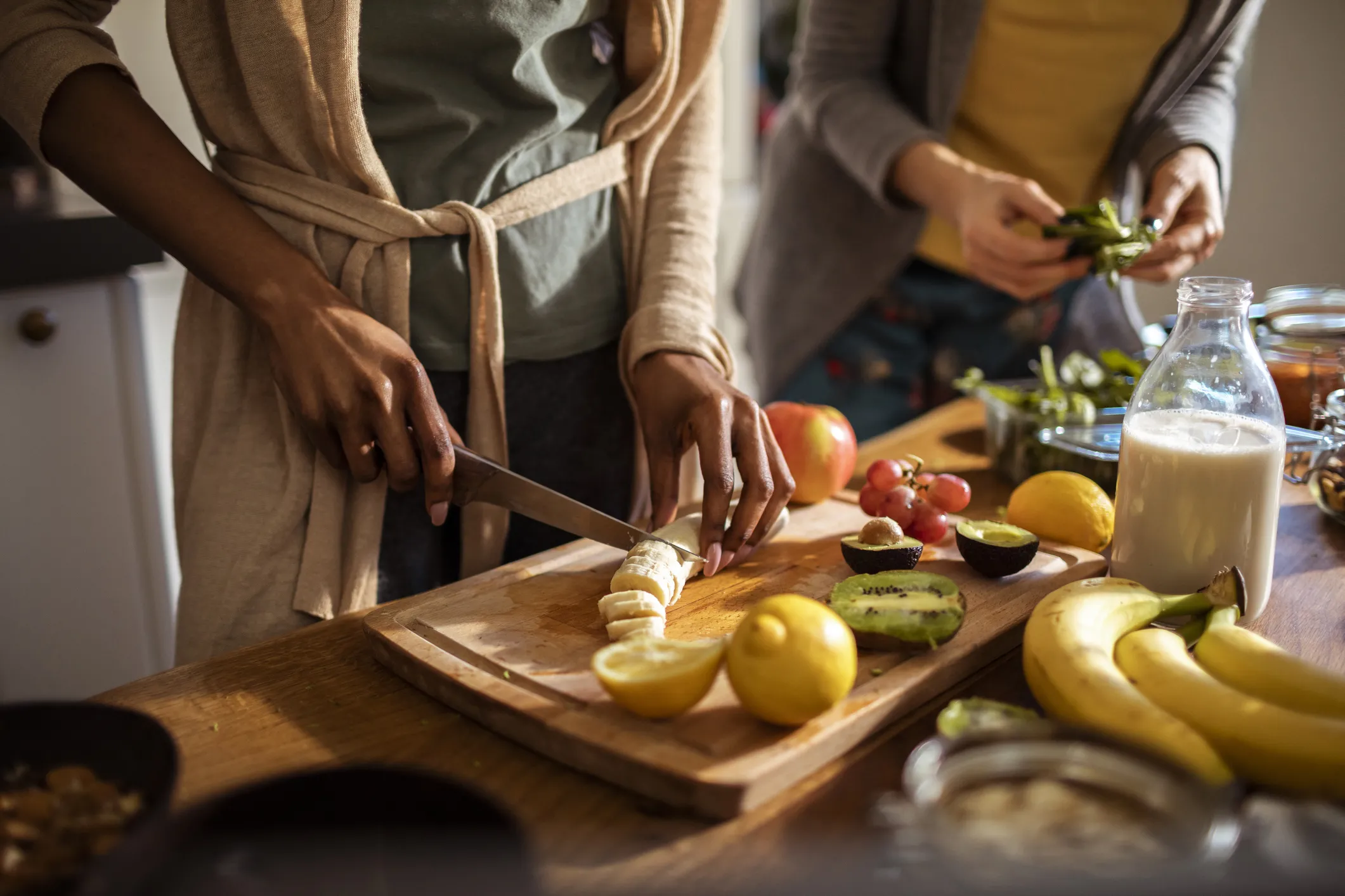 Women prepare veggies and fruits on a cutting board in a brightly lit kitchen