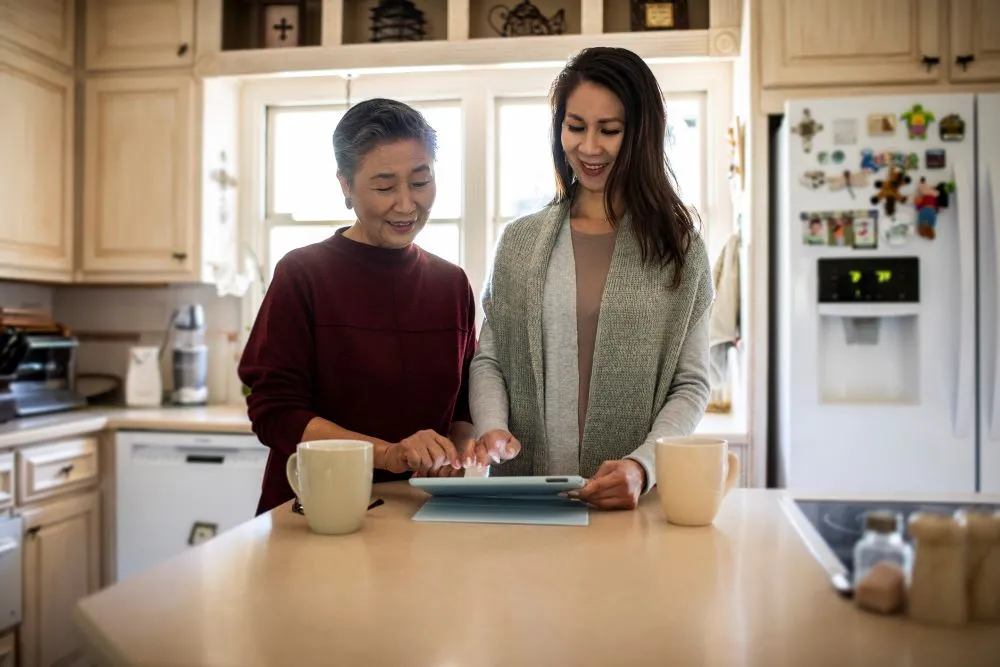 A mother and daughter smile while looking at a digital tablet on their kitchen table