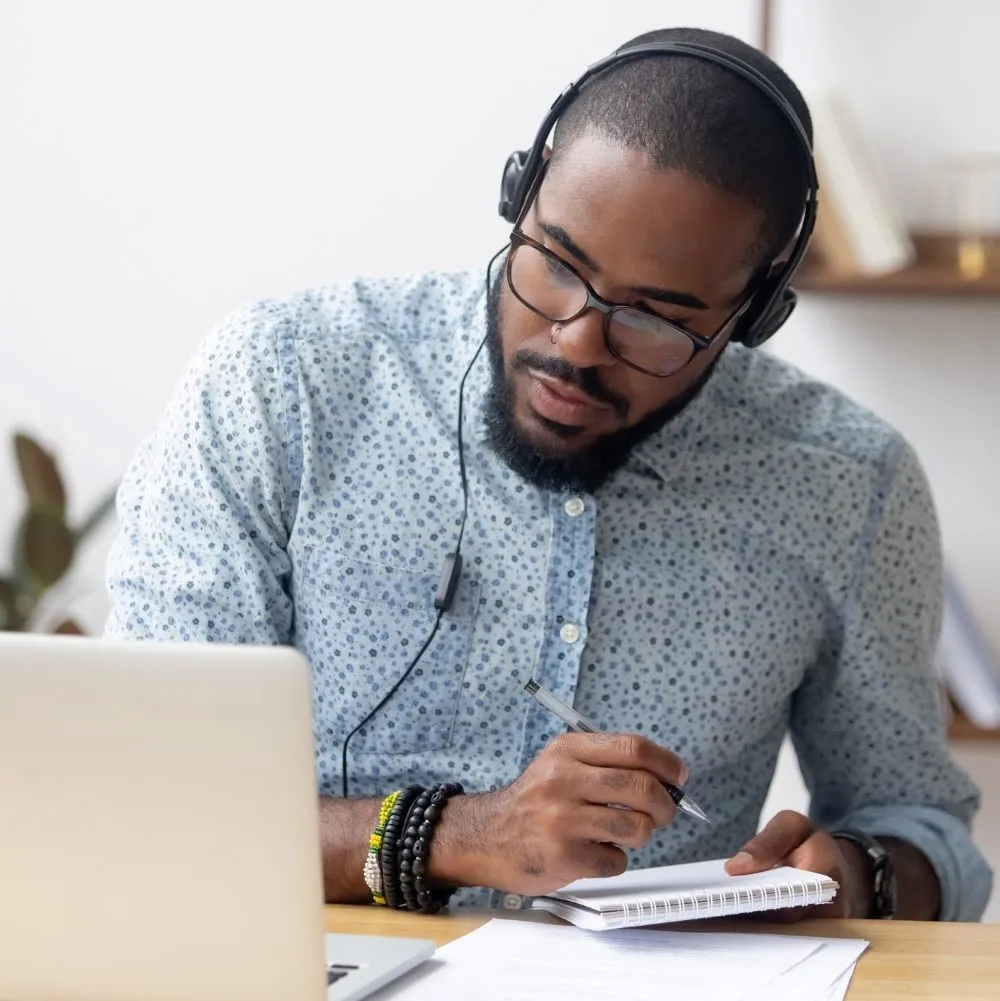 A man wearing headphones views his laptop screen and takes notes with a pen in his hand