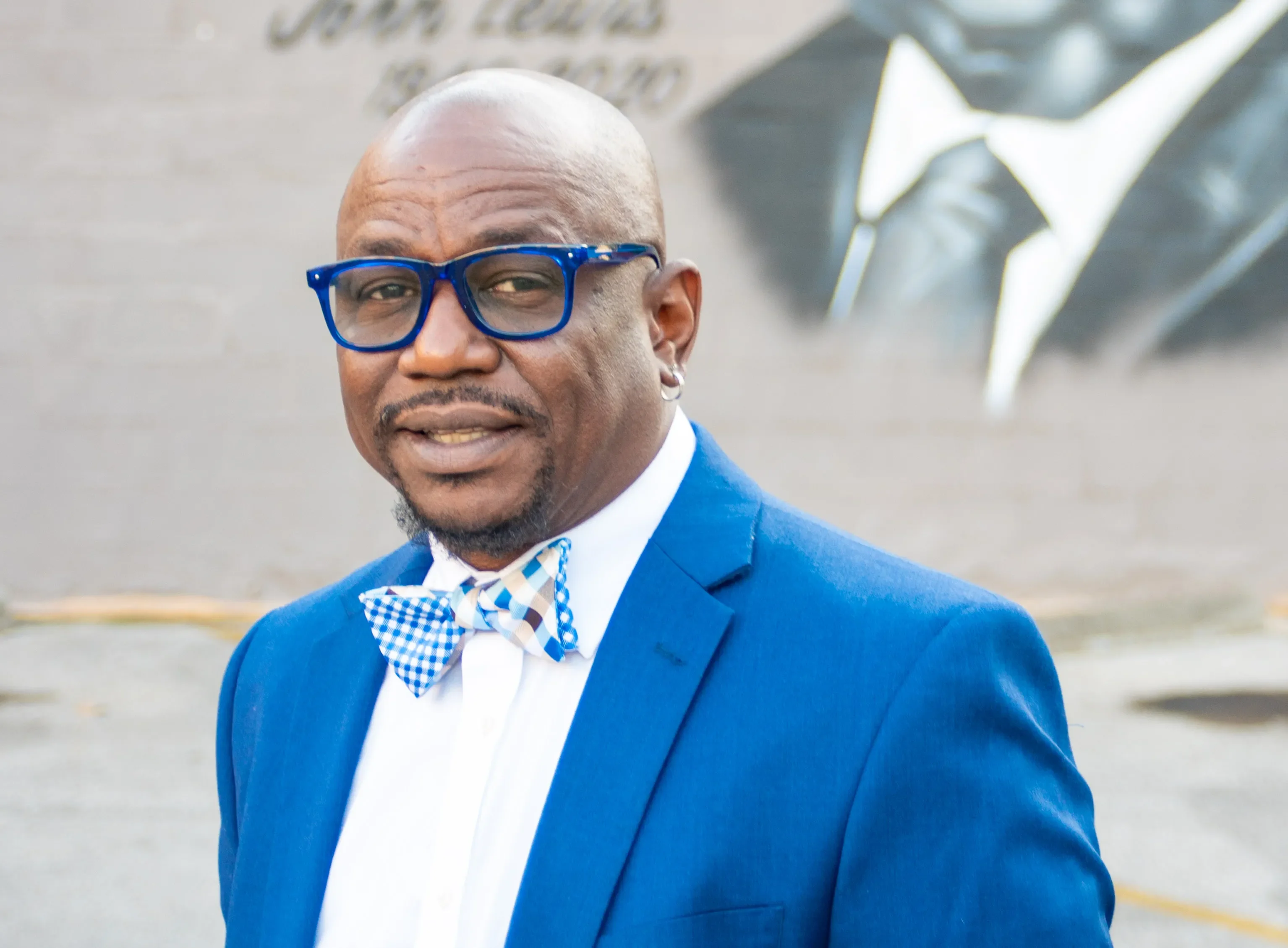Tony Walker wears a blue and white bow tie, blue blazer, white blouse, and glasses with blue frames